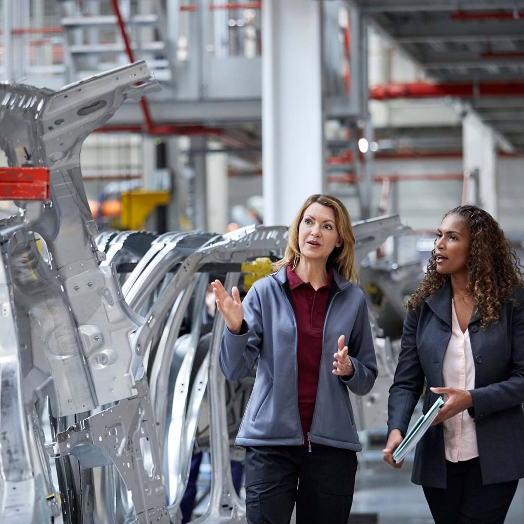 Two women walking floor of auto manufacturer looking at assembly line